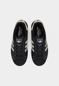 Ζευγάρι μαύρα αθλητικά παπούτσια Adidas με μπεζ ρίγες και σχεδιασμό με μύτη σε σχήμα κοχύλι, οπτική από πάνω σε ανοιχτό φόντο.