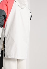 Veste à capuche blanche et rouge avec accents noirs, présentant une coupe décontractée, une texture lisse et des fermetures éclair latérales pour la ventilation.