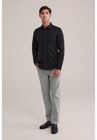 Juoda marškiniai su ilgomis rankovėmis, derinami su šviesiai pilkomis tekstūruotomis kelnėmis ir juodais slip-on batais. Paprasta fonas, be raštų.