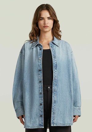 Frau mit schulterlangem braunem Haar trägt eine übergroße hellblaue Jeansjacke über einem schwarzen Oberteil und schwarzen Hosen vor einfachem Hintergrund.