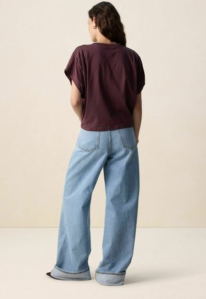 Bordeauxrood kort T-shirt met opgerolde mouwen, gecombineerd met lichtblauwe wijde jeans met omgeslagen zoom en achterzakken. Effen achtergrond.