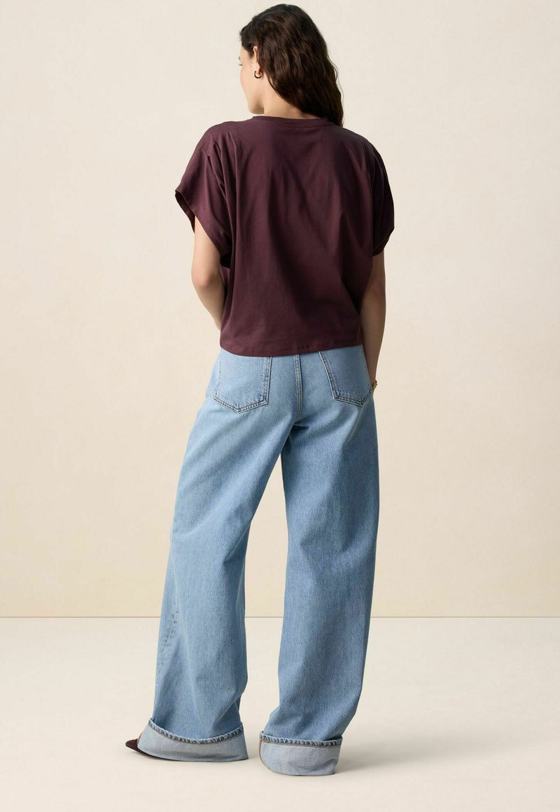 Bordeauxrood kort T-shirt met opgerolde mouwen, gecombineerd met lichtblauwe wijde jeans met omgeslagen zoom en achterzakken. Effen achtergrond.