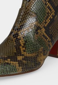 Gros plan d’une chaussure à talon pointu recouverte d’un motif en cuir de serpent brillant vert, marron et noir avec un talon bloc rouge.