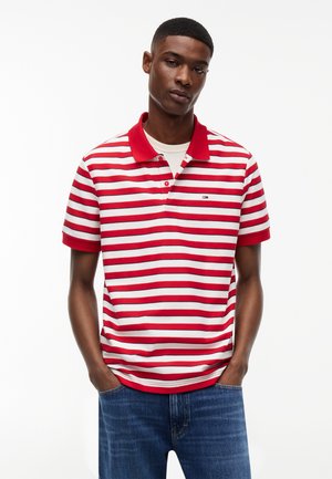 Jeune homme portant un polo à manches courtes rayé rouge et blanc avec col, associé à un jean bleu, debout les mains dans les poches.