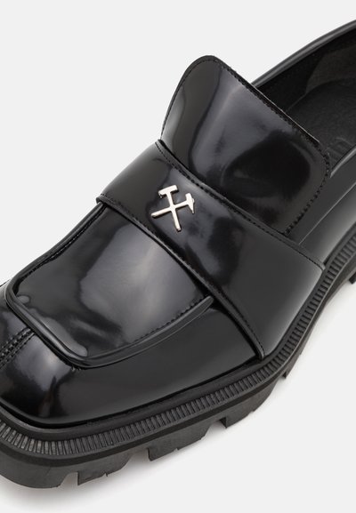 Black patent leather loafer with a thick rubber sole. Features a wide strap and metallic X emblem on the upper. Smooth, glossy finish.