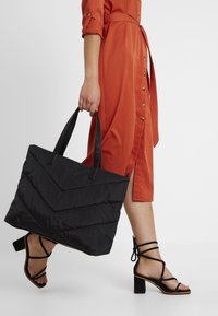 Femme en robe orange marchant, portant un grand sac fourre-tout noir matelassé, chaussée de sandales à talons blocs noires avec des bretelles.