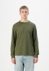 Ung mann med kort blondt hår som har på seg en olivengrønn langermet skjorte med ribbestrikk og lyseblå jeans mot en hvit bakgrunn.
