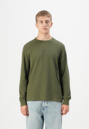 Ung man med kort blont hår iförd en ribbad olivgrön långärmad tröja och ljusblå jeans mot en vit bakgrund.