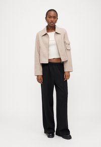 Beige kort jacka med framdragkedja, en bröstficka och en texturerad yta, kombinerad med svarta vida byxor och svarta skor.