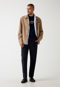 Giacca beige con bottoni sopra una t-shirt blu navy con logo, abbinata a pantaloni blu navy e mocassini neri. Design minimalista con colori a contrasto.