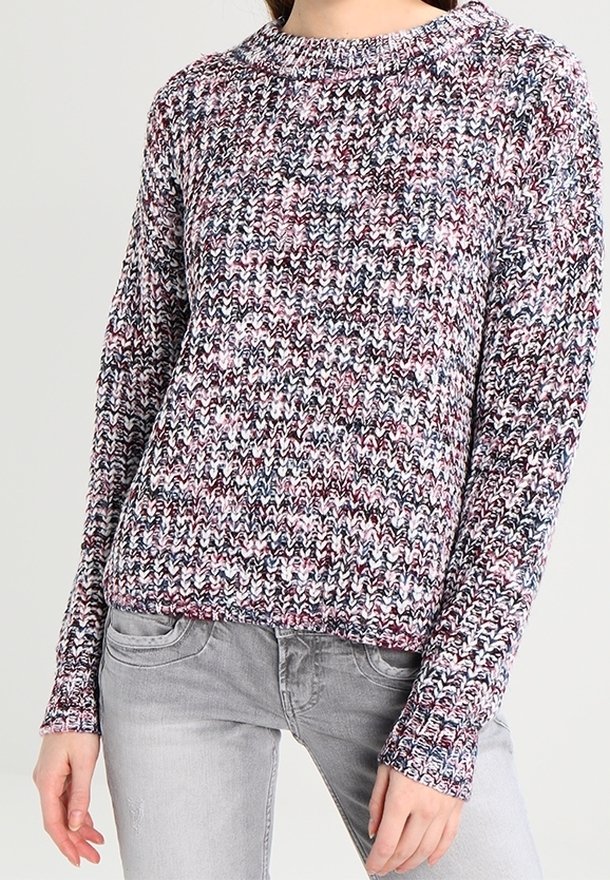 Žena nosící vícebarevný pletený svetr s kulatým výstřihem a dlouhými rukávy, spárovaný s světle šedými džíny.