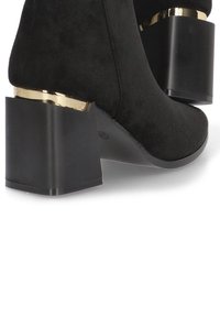 Botín de ante negro hasta el tobillo con un tacón grueso negro y un acento dorado en la base. Textura suave con una forma redondeada para un diseño moderno.