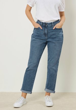 Donna in piedi con jeans blu risvoltati, top bianco a maniche corte con occhielli e sneakers bianche, mani in tasca su sfondo neutro.