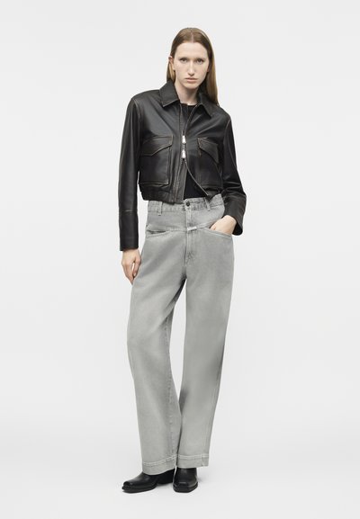 Woman wearing a black cropped leather jacket, loose light gray high-waisted jeans, and black ankle boots, standing with hand in pocket.