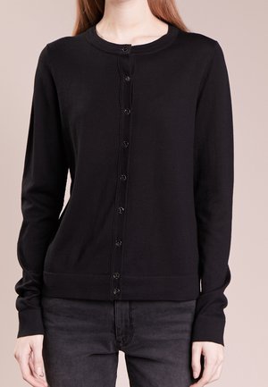 Femme portant un cardigan noir boutonné à manches longues avec un col rond et un pantalon foncé sur un fond uni.