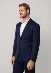 Granatowa marynarka z gładkiego materiału, zapięcie na pojedynczy guzik, klapy w stylu notch oraz dwoma przednimi kieszeniami, zestawiona z białym polo poniżej.