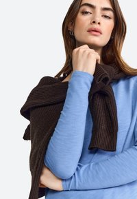 Brown knitted sweater draped over a light blue long-sleeve top. The sweater has ribbed texture, while the top is smooth and fitted.