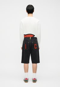 Άτομο με την πλάτη γυρισμένη, φοράει λευκό πουκάμισο με μακριά μανίκια, μαύρα σορτς μέχρι το γόνατο με κόκκινα γράμματα "I" και "C" στις πίσω τσέπες, λευκές κάλτσες και πολύχρωμα παπούτσια.