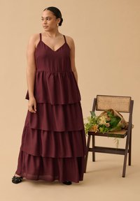 Femme en robe maxi bordeaux sans manches à volants, debout près d'une chaise en bois, tenant un bouquet de fleurs mêlées enveloppé dans du papier.