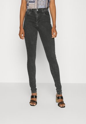 Kobieta ubrana w czarne skinny jeans z wysokim stanem, wyblakłe, z wzorzystym bezrękawnikiem i czarne sandały na obcasie, stojąca na jednolitym tle.