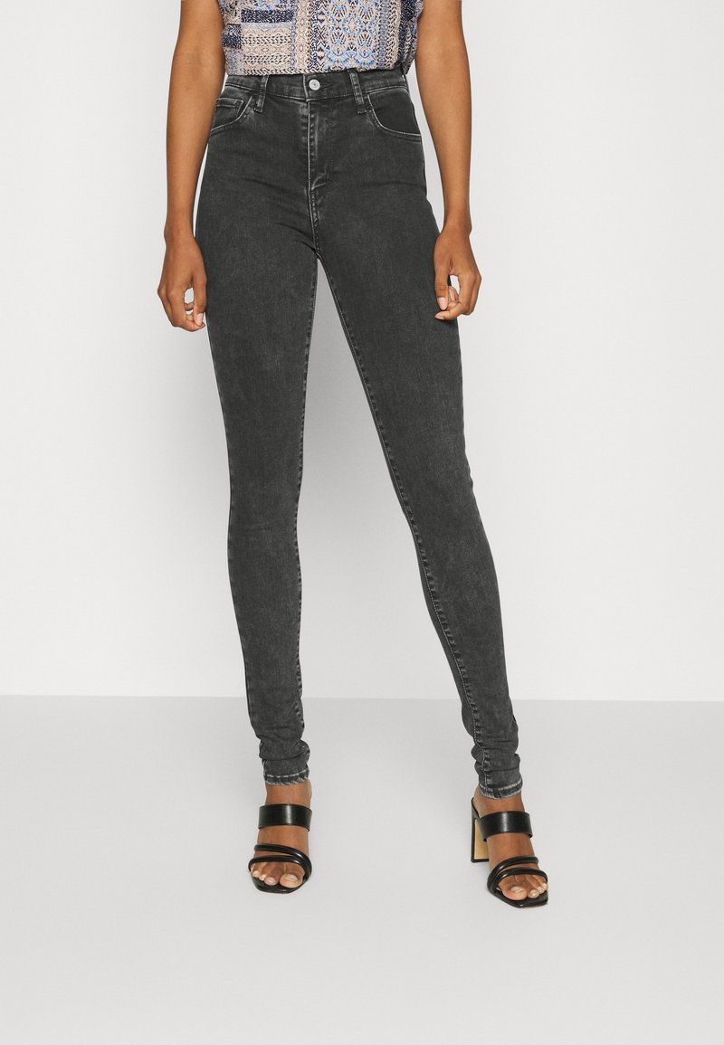 Femme portant un jean skinny noir délavé taille haute avec un top sans manches à motifs et des sandales noires à talons, debout devant un fond uni.