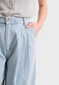 Pantalones de mezclilla de pierna ancha en azul claro, con cierre de botón y pliegues, que cuentan con bolsillos laterales y una textura suave.
