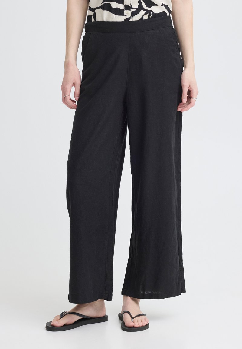 Personne portant un pantalon noir ample, des tongs noires et un haut à motifs, debout devant un fond uni.