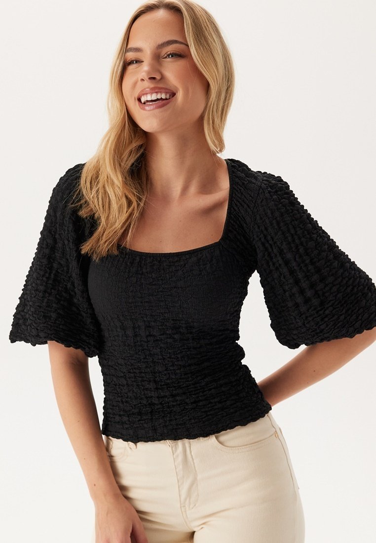 Top nero arricciato con scollatura quadrata, maniche corte voluminose e finitura testurizzata, indossato con pantaloni chiari.