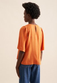 Top in lino arancione con maniche corte a sbuffo e dettaglio a 'keyhole' sulla schiena, abbinato a jeans in denim blu. Tessuto morbido e vestibilità rilassata.