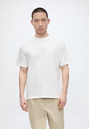 Młody mężczyzna ubrany w gładki biały t-shirt z krótkim rękawem i beżowe spodnie, stojący na jednolitym białym tle.