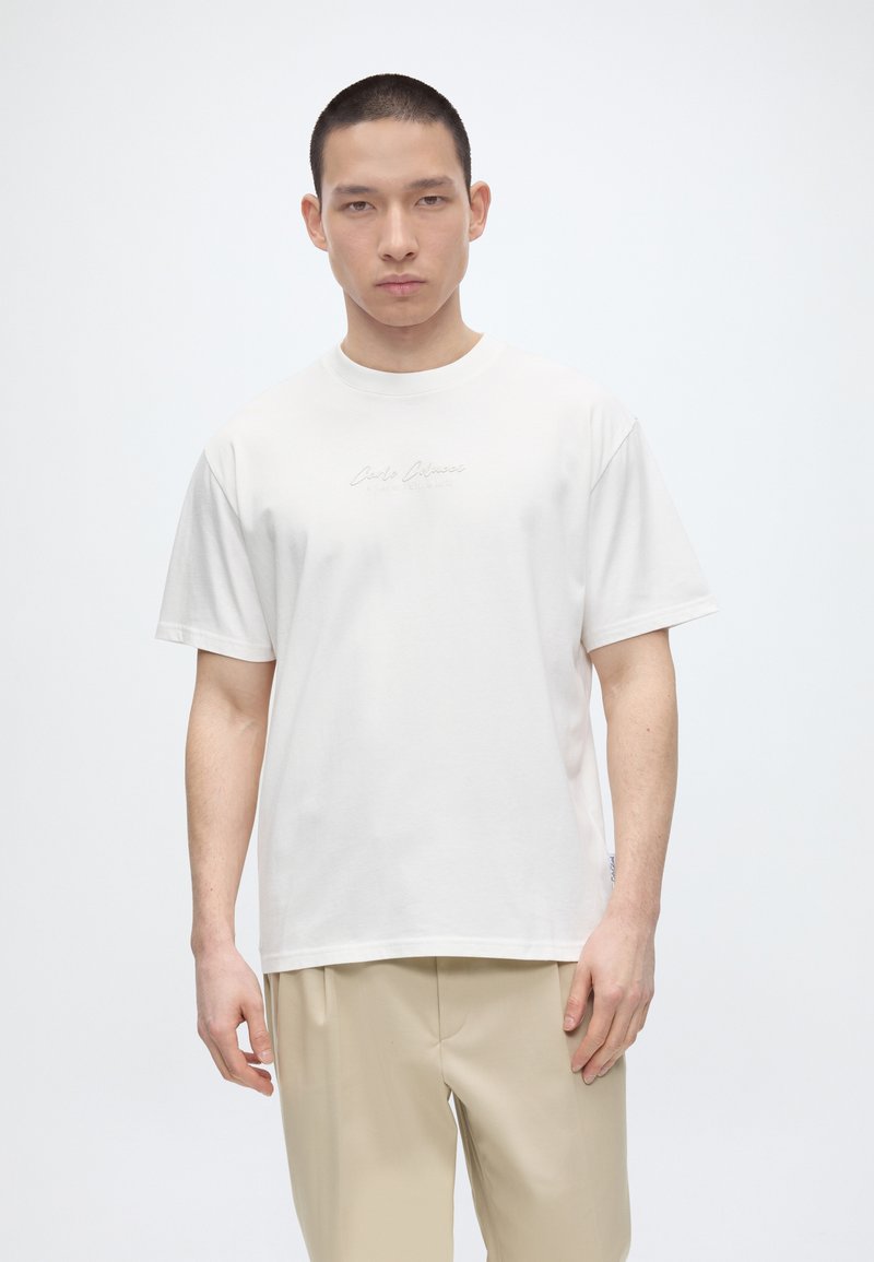 Νέος άνδρας που φοράει απλό λευκό κοντομάνικο T-shirt και μπεζ παντελόνι, στέκεται μπροστά σε απλό λευκό φόντο.