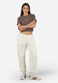 T-shirt corta marrone con lettering bianco, pantaloni della tuta color crema con vita elastica e tasche laterali, abbinati a sneakers bianche con strisce marroni.