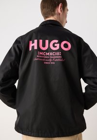 Črna jakna z velikim roza logotipom "HUGO" na hrbtu, gladka tekstura in sproščen dizajn s straight hem in dolgimi rokavi.