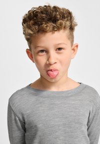 Graues Langarmshirt mit einem entspannten Ausschnitt; Kind mit hellbraunen lockigen Haaren, das die Zunge rausstreckt und einen verspielten Gesichtsausdruck zeigt.
