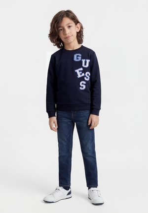 Garçon aux cheveux bruns mi-longs portant un pull bleu marine avec les lettres "GUESS", un jean bleu et des baskets blanches, debout sur un fond blanc.