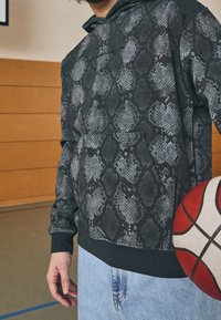 Felpa con cappuccio nera e grigia con un motivo in pelle di serpente, dotata di tasca frontale e polsini a coste, abbinata a jeans blu chiaro e a un pallone da basket.