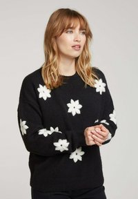 Pull noir avec application florale blanche. Texture douce, coupe ample, finitions côtelées aux poignets et à l'ourlet. Col rond, avec plusieurs motifs floraux sur l'ensemble.