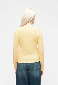 Γυναίκα με μακριά ξανθά μαλλιά, που φορά φωτεινό κίτρινο ριμπ πλεκτό μακρυμάνικο τοπ και μπλε τζιν, με πλάτη σε απλό φόντο.