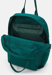 Sac à dos en toile teal avec fermeture à glissière, grand compartiment principal, poche zippée interne et deux poignées supérieures. Texture lisse, forme structurée.