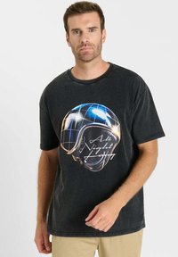 Zwart katoenen T-shirt met een afbeelding van een helm met een blauw rasterontwerp en de tekst "All Night Long" in het wit. Korte mouwen, ontspannen pasvorm.