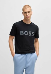 T-shirt noir à manches courtes arborant le logo "BOSS" en gris texturé. Design à col rond avec un tissu doux. Associé à un pantalon bleu clair.