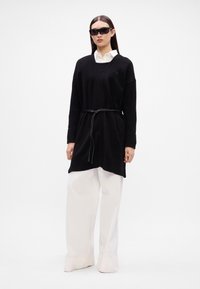 Μαύρο πλεκτό cardigan με V-λαιμόκοψη, μακριά μανίκια και ζώνη στη μέση. Συνδυάζεται με φαρδιά λευκά παντελόνια και μαύρα γυαλιά ηλίου.