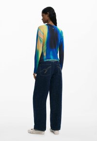 Long-sleeve shirt with a blue, green, and yellow abstract design; paired with wide, dark blue denim jeans featuring golden stitching.