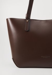 Sac tote en cuir marron avec une texture lisse, une large ouverture et une seule sangle. Présente un accent doré et un logo embossé à l'avant.