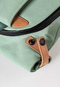Groene canvas tas met een gladde textuur, leren accenten, zwarte rits en een flap die omgevouwen kan worden. Bijzonderheden zijn een opvallende drukknoopsluiting en stikseldetails.