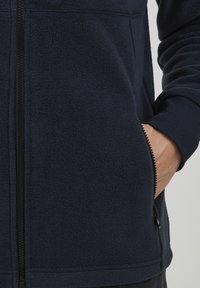 Marineblå fleecejakke med glidelås foran, ribbestrikkede mansjetter og sidelommer. Teksturen er myk og litt strukturert, noe som gir varme.