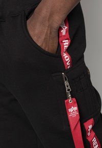 Svarta cargobyxor har en sidficka med dragkedja, röd "REMOVE" remdetail och texturerade sömmar för en funktionell design.