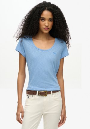 Lyseblå t-shirt med korte ærmer og scoop-hals samt diskret logo. Båret over hvide bukser, fremhævet af et brunt bælte. Blødt stof.