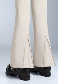 Beżowe spodnie w kształcie dzwonów z zamkami błyskawicznymi na dole, wykonane z gładkiego materiału, w połączeniu z wytrzymałym czarnym obuwiem.