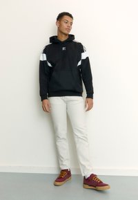 Felpa sportiva nera con dettagli bianchi, tasca a marsupio, abbinata a jeans bianchi e sneaker bordeaux con lacci gialli e suola in gomma.